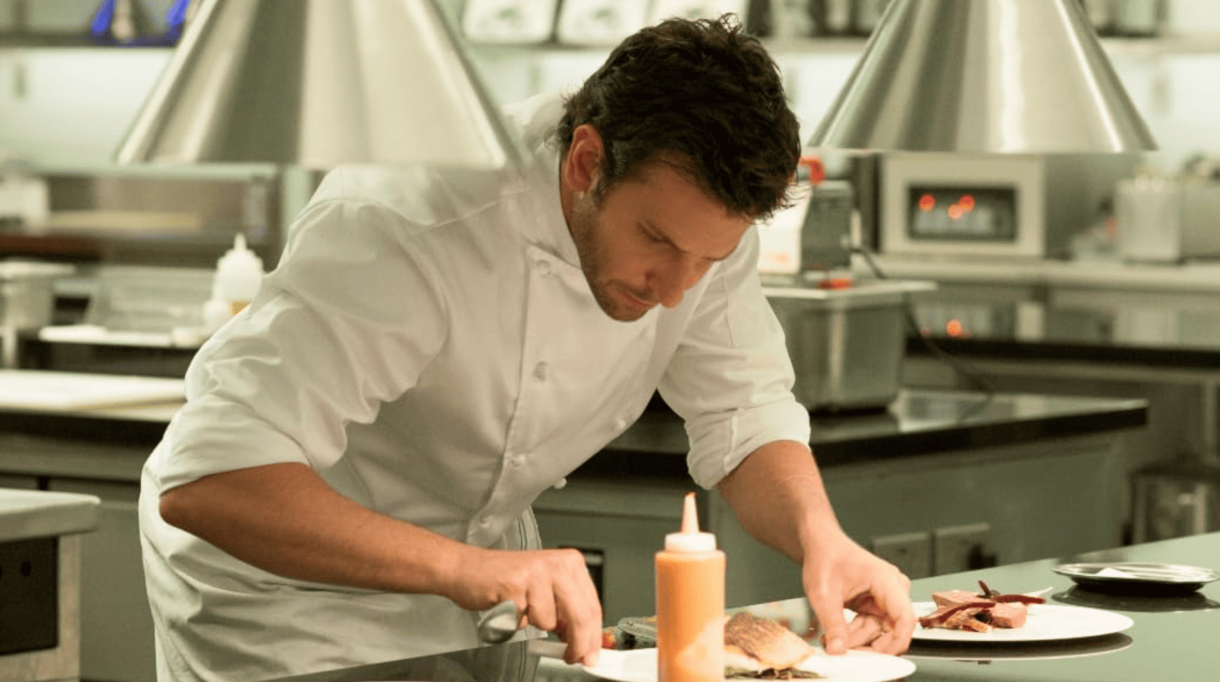 Bradley Cooper dressed in a chef uniform standing in an industrial kitchen putting the finishing touches on a dish in this image from 3 Arts Entertainment.