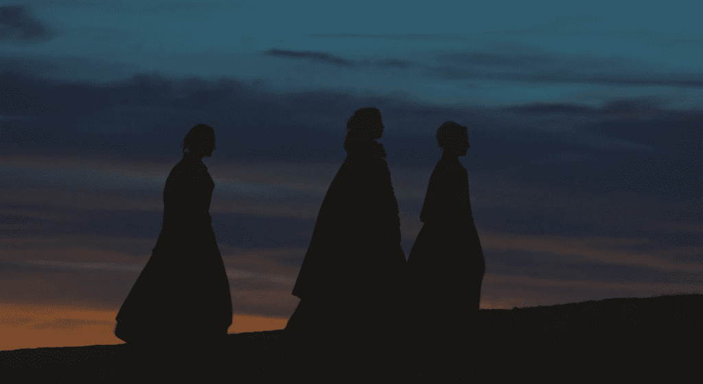 Three silhouettes of hooded, caped figures walking up a hill at sunset