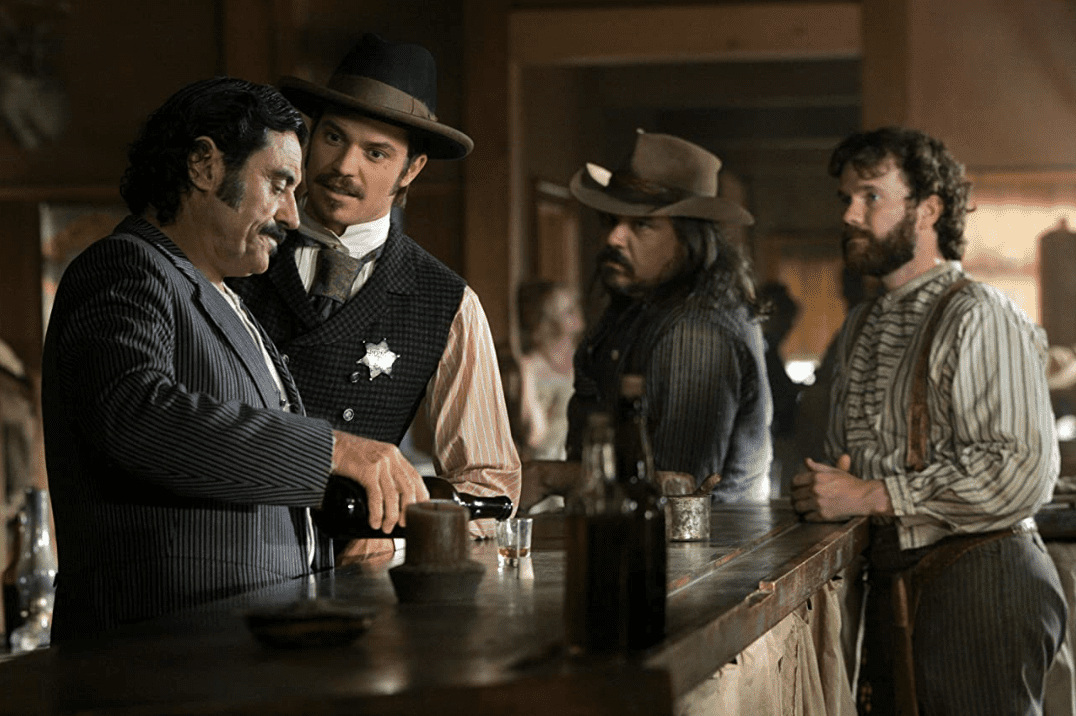 Deadwood cast standing around a bar counter