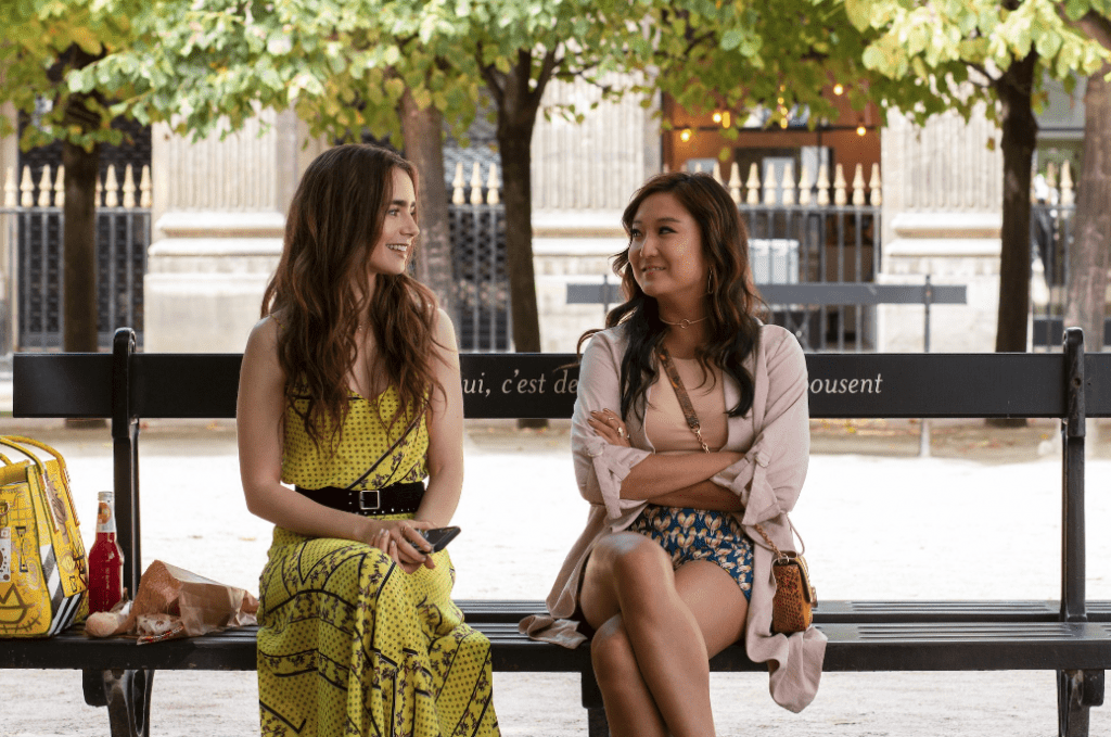 Emily and Mindy sit on a park bench, swapping stories of their first experiences in Paris. 