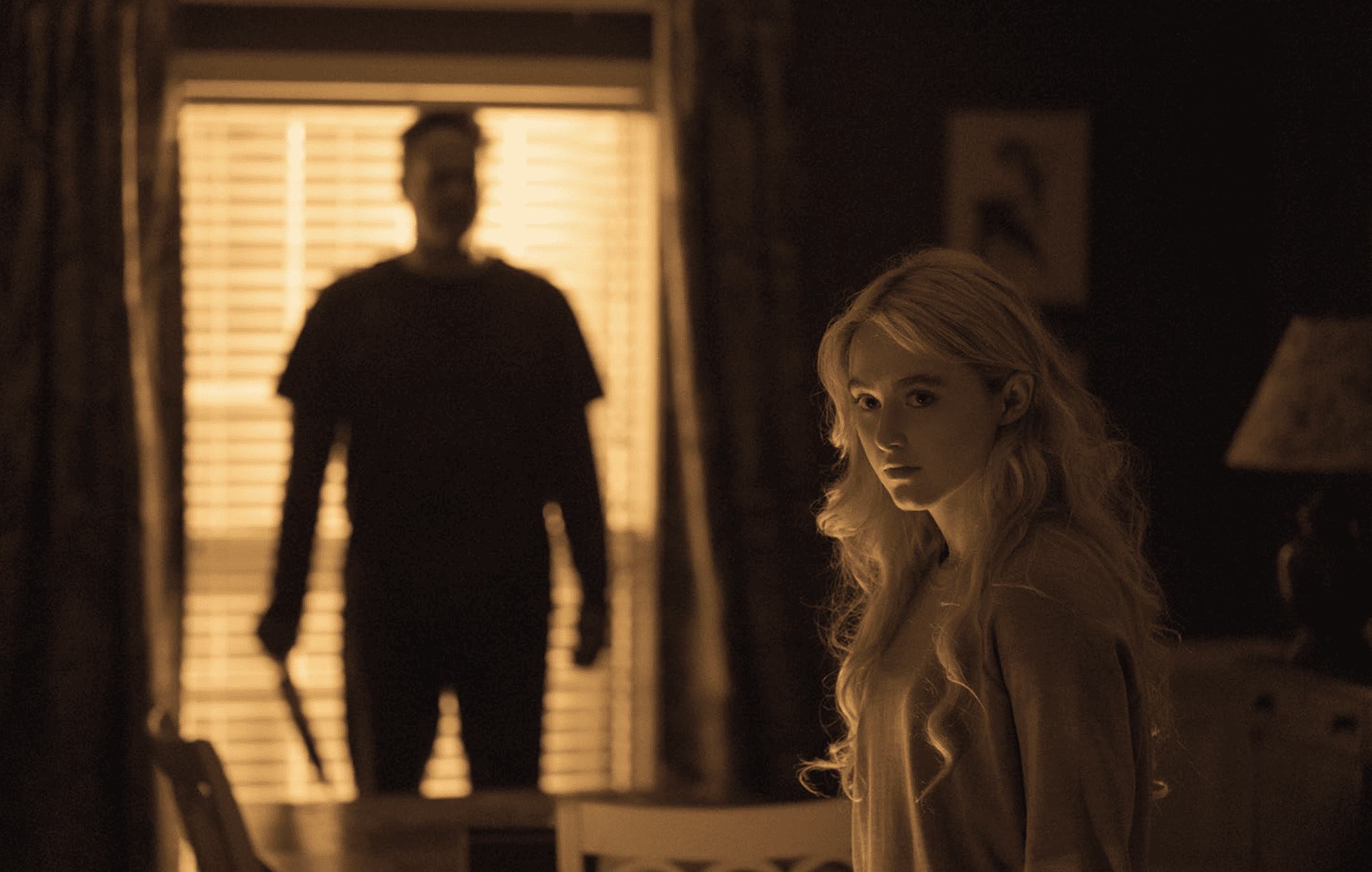 A girl looks into the camera as a silhouetted man with a knife watches her in this image from Blumhouse Productions.