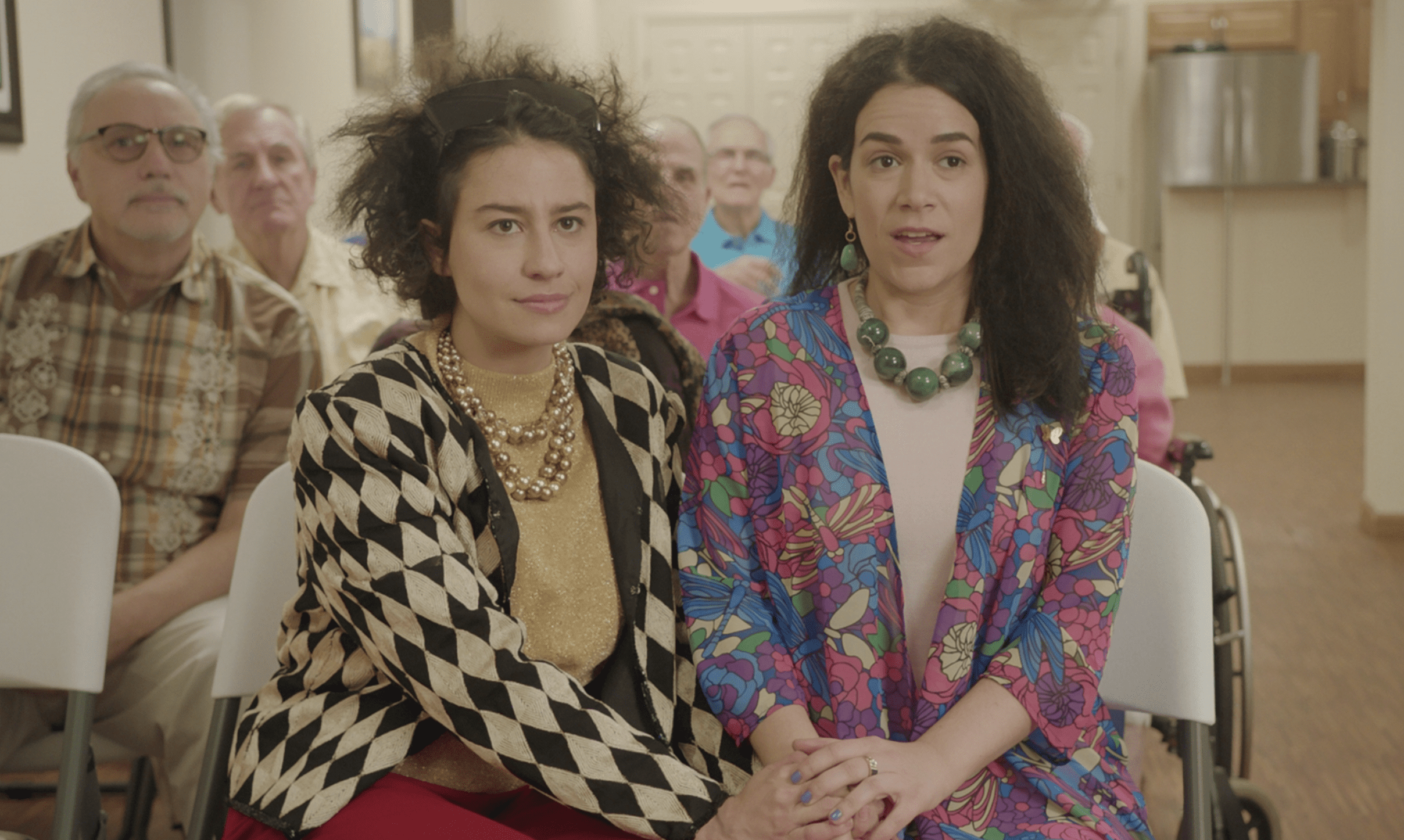 The two main characters of Broad City sit together, holding hands and wearing clothes meant for seniors.