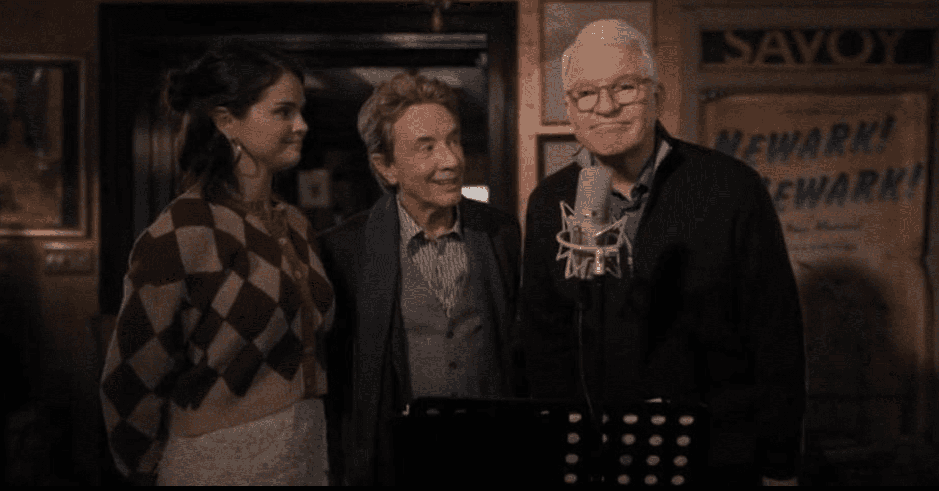 Steve Martin talks into a microphone as Selena Gomez and Martin Short observe closely.