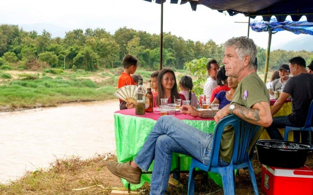 Anthony Bourdain sitting near a river in Laos and sharing a meal with locals