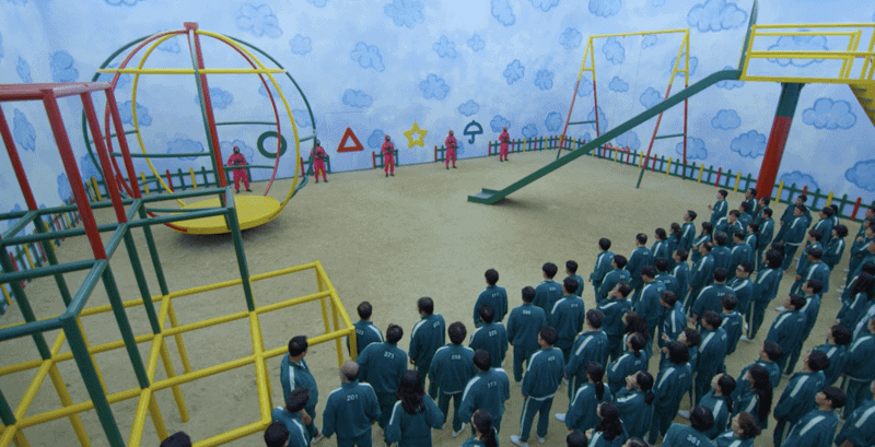 A group of people wearing matching, numbered jumpsuits wait in a room decorated with crayon-like clouds and a jungle gym.