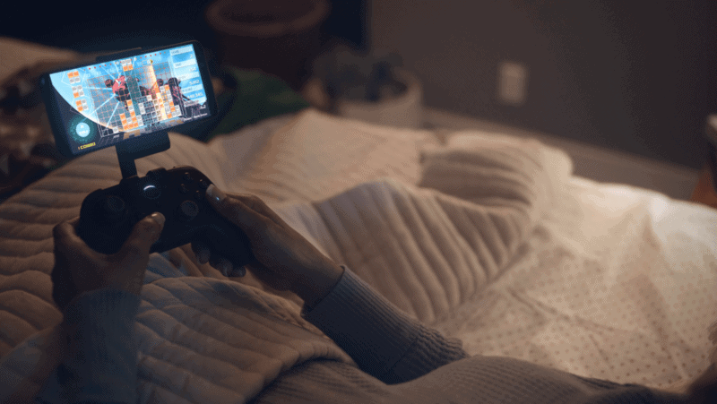 A person in bed playing games via an Amazon Luna controller with a smartphone attached