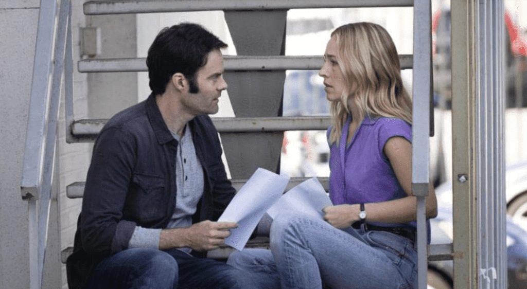 A man in a dark blue shirt and jeans and a woman in a lavender shirt and jeans sit facing each other on a fire escape while holding scripts.