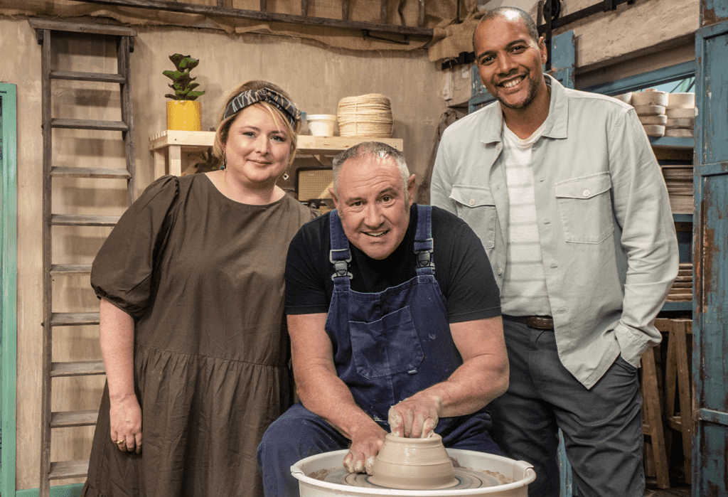 The host and judges of the show, one of whom is seated at a throwing wheel, pose for a photo in the studio.