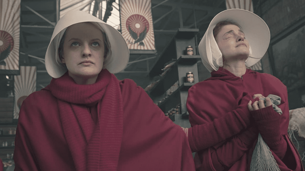 Handmaids June and Janine in red dresses holding each other’s hands with fear and anticipation.