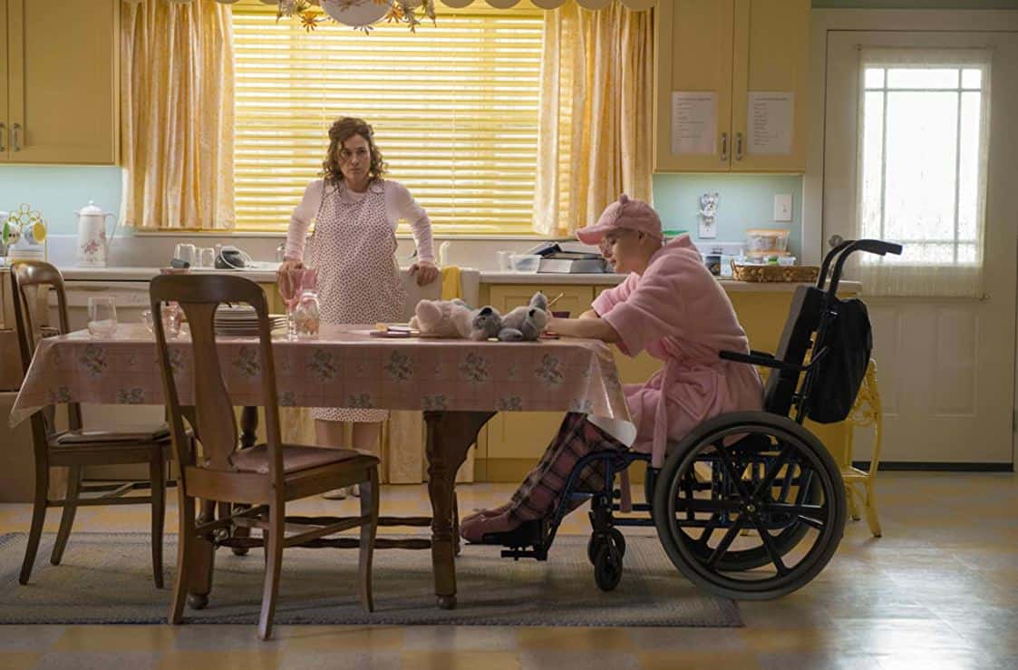 Dee Dee Blanchard stands by the kitchen sink and looks at Gypsy Rose Blanchard at the table in the show “The Act”