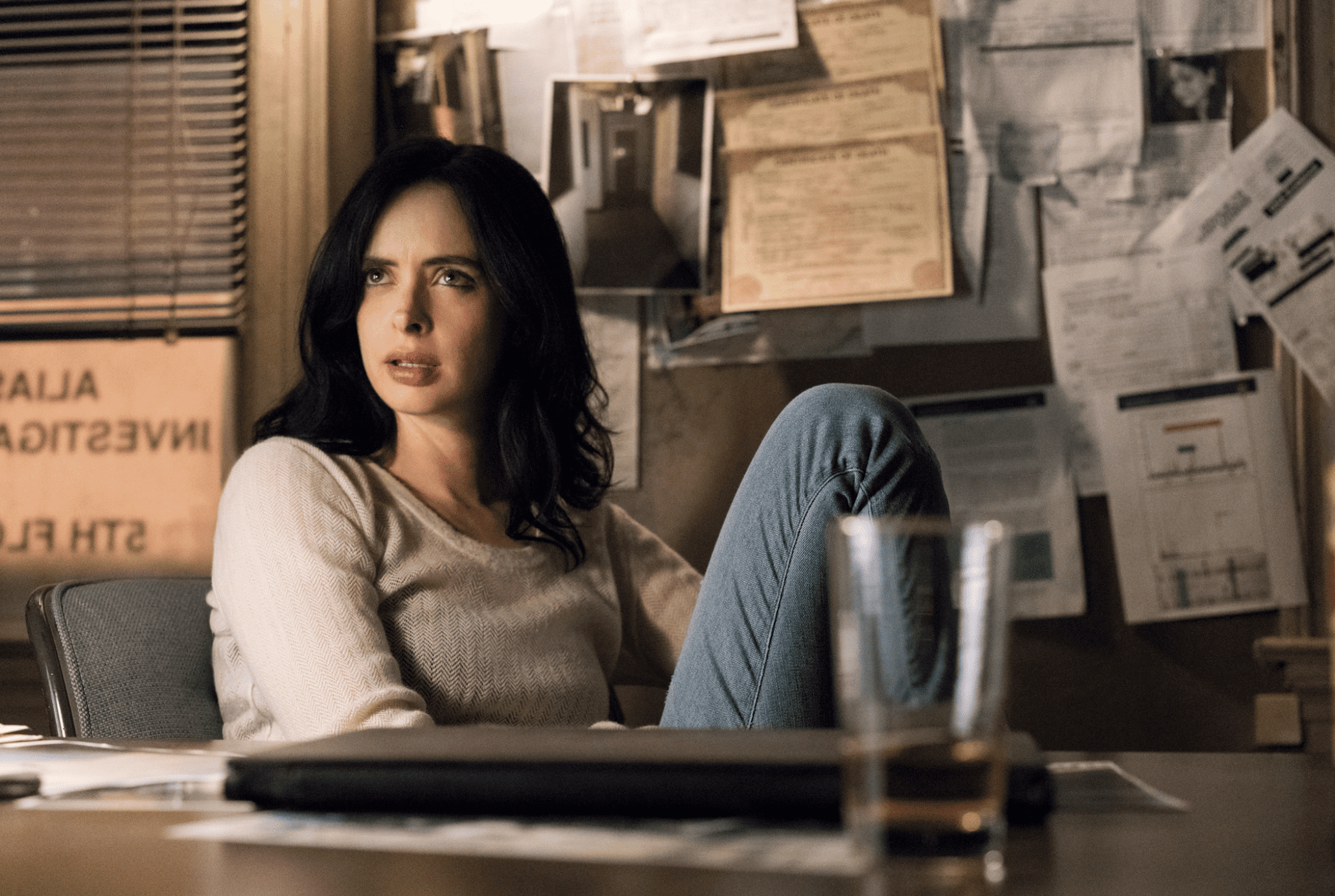 A woman lounges at a desk with the wall behind her covered in papers in this image from 3 Arts Entertainment. 