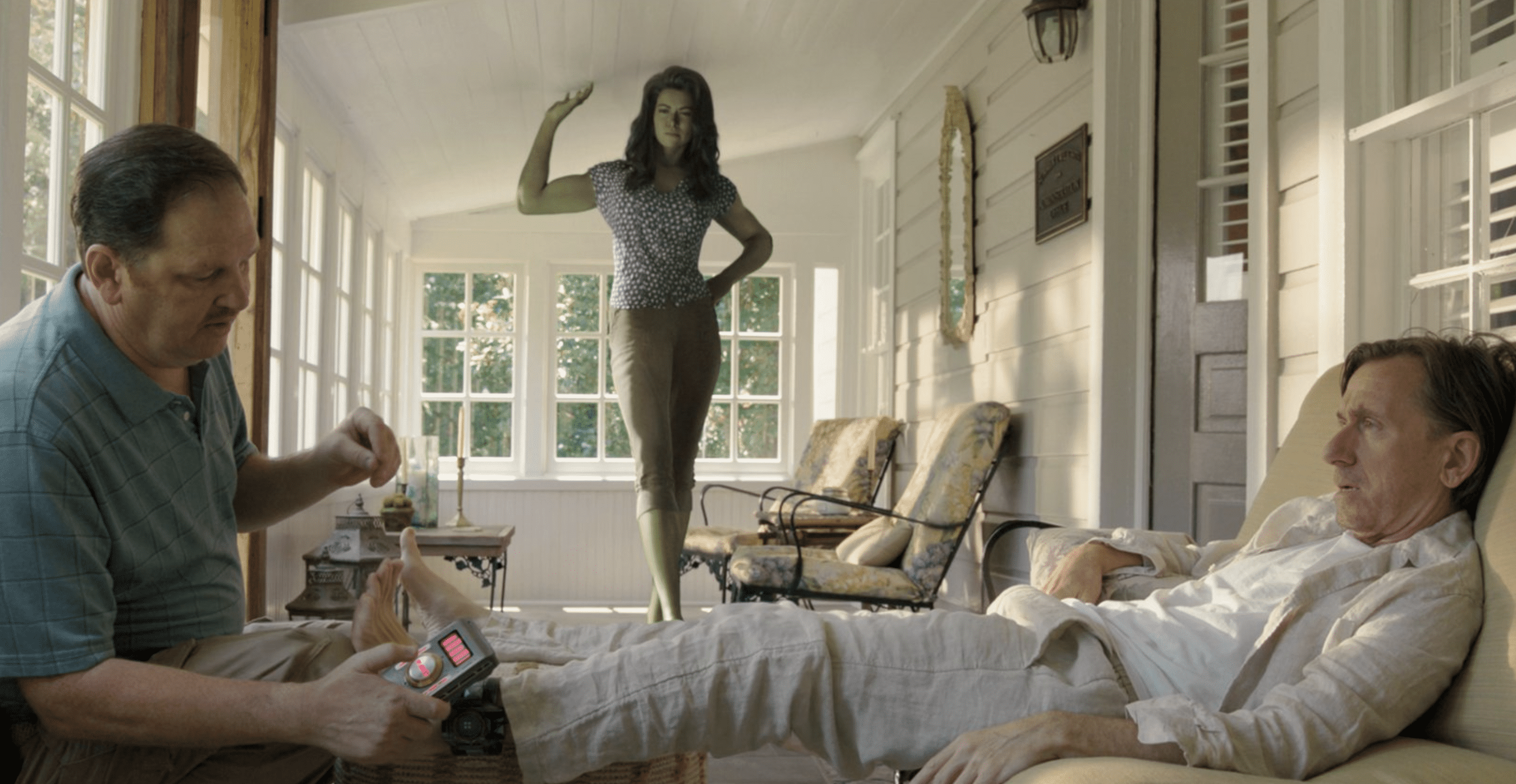 She-Hulk, a giant green superhuman, stands in a sunny room while a lounging man in linen clothes gets his ankle monitor checked.
