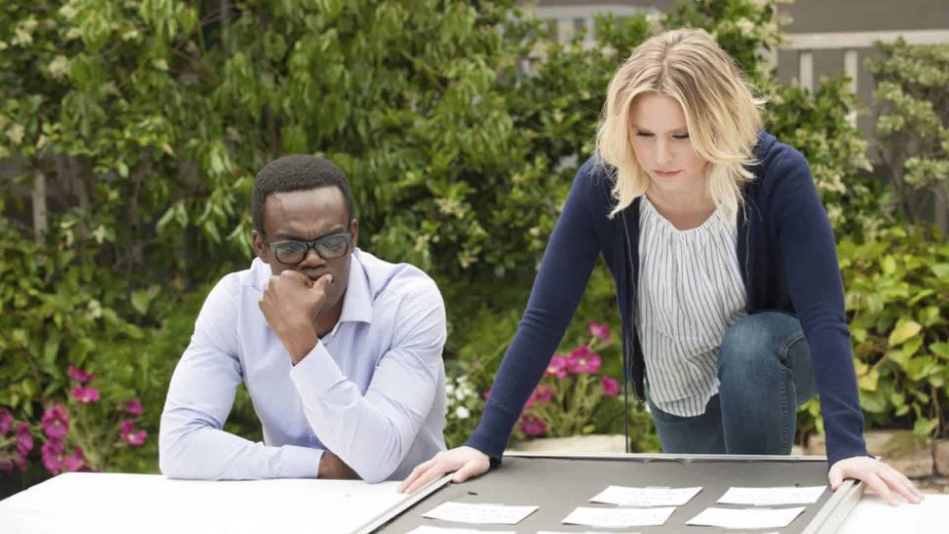 Chidi and Eleanor examine a table with cards on it.