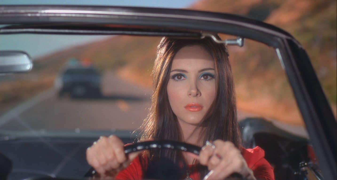 Elaine Parks in a red dress driving a convertible in front of a police car