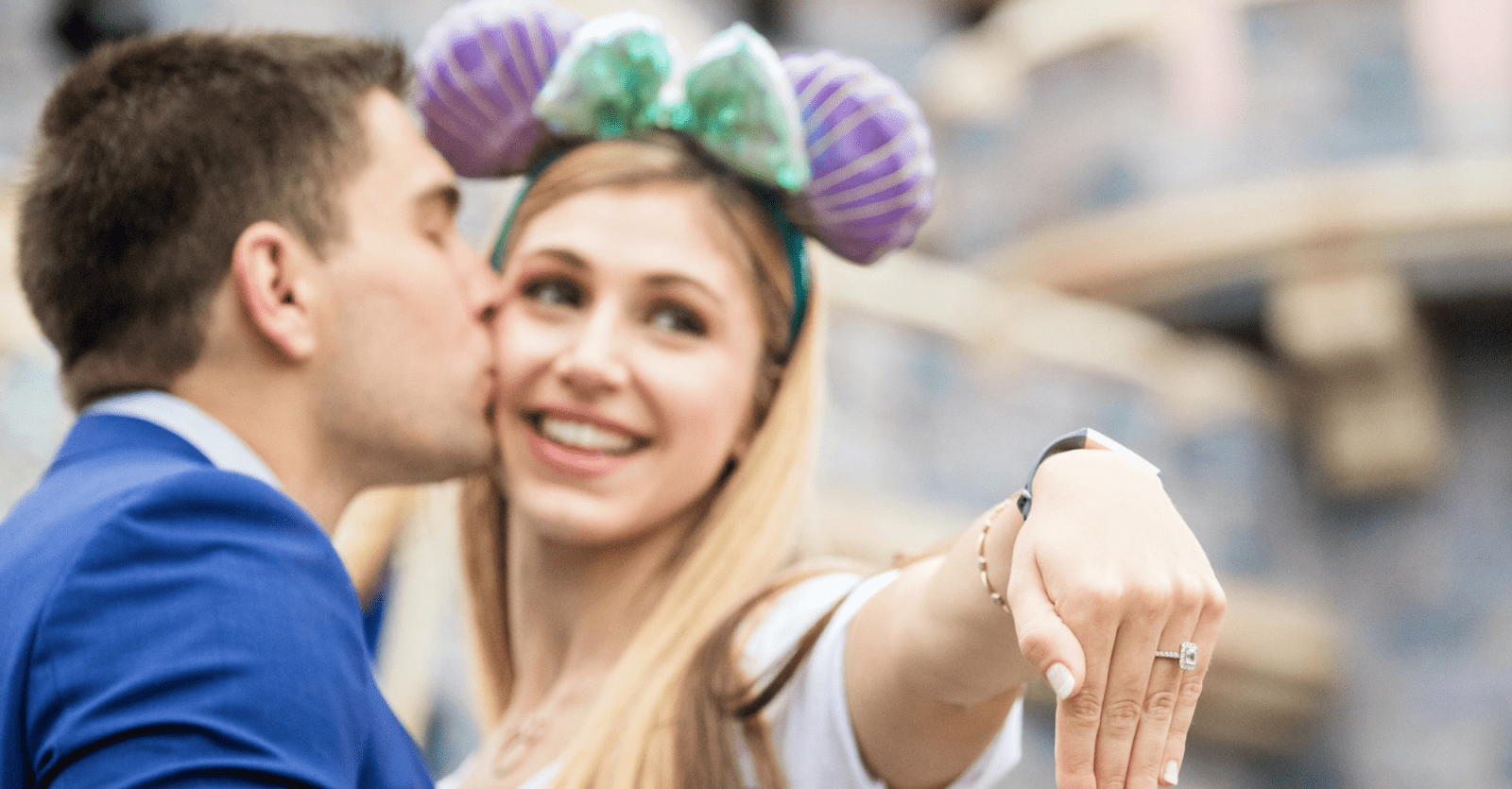 A bride-to-be holding out her hand, showcasing her wedding ring while her fiancé kisses her cheek