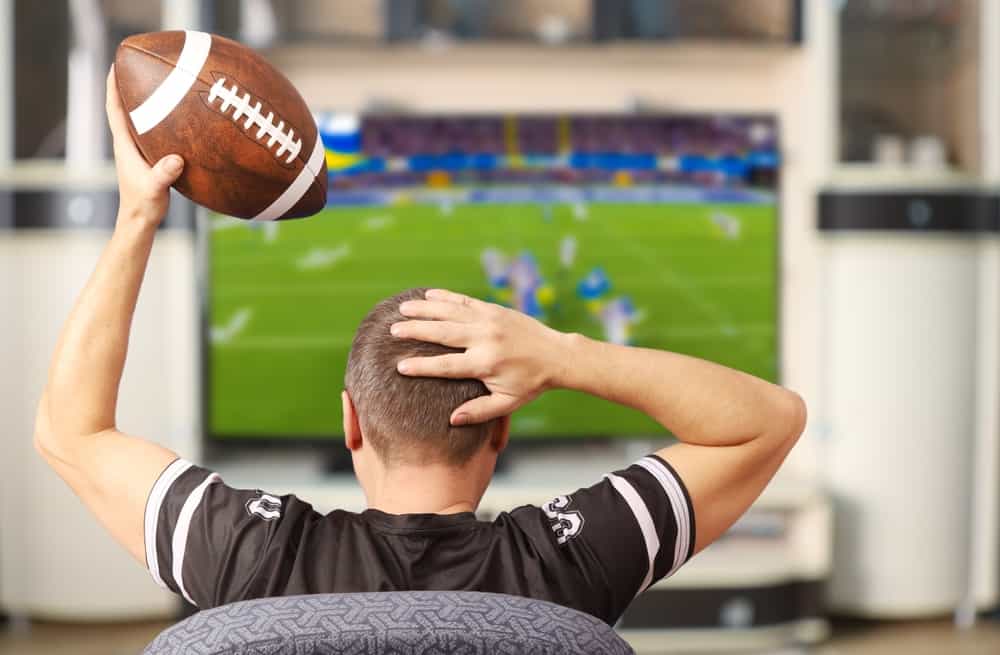 A man holds a football in one hand while watching a game on a blurred TV