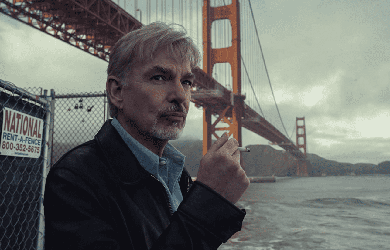 A pensive man standing under the Golden Gate Bridge looks off camera while holding a cigarette. 