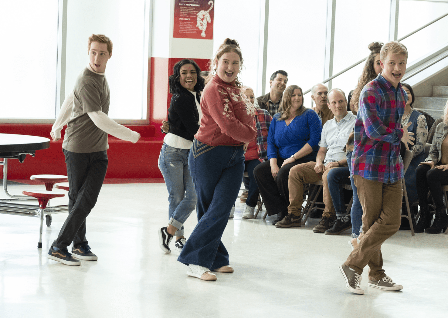 A group of students dances in the middle of what seems to be a high school cafeteria.