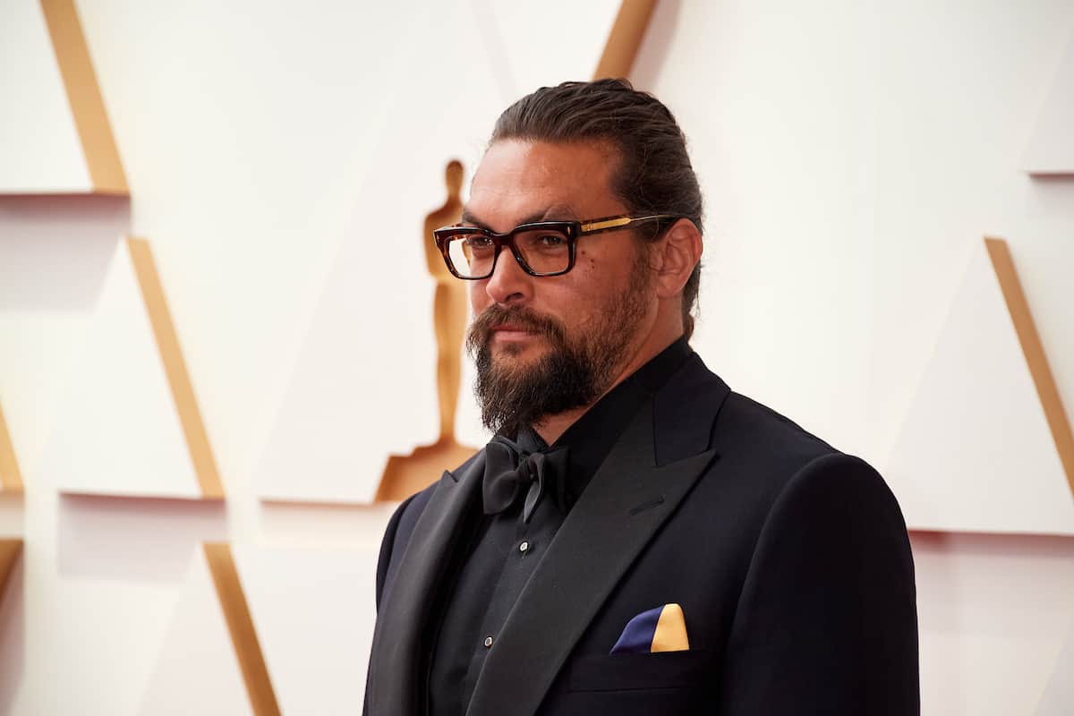 Jason Momoa standing in front of the Oscars logo on the red carpet