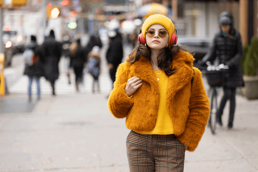 Mabel in a fashionable outfit walking an NYC street with headphones on