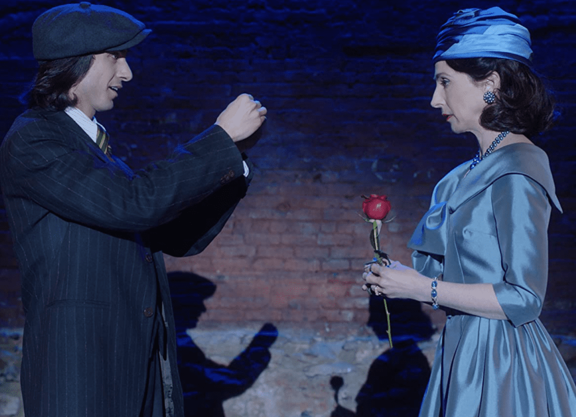 A woman in profile with an elegant dress and hat stands on a stage holding a rose and staring at a man holding up his hands