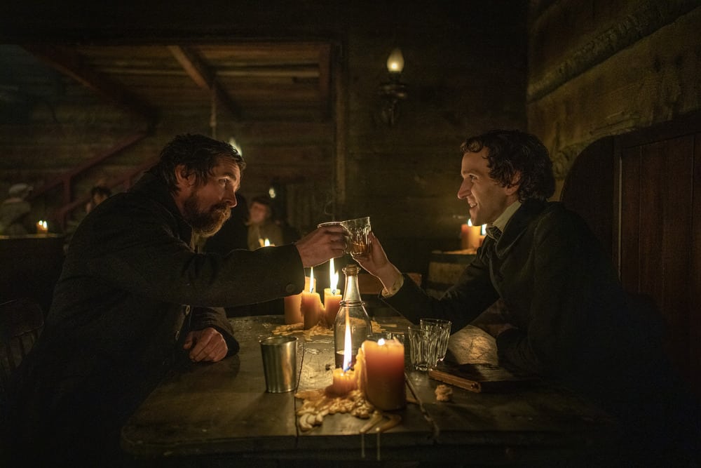 Augustus and Edgar clink glasses by candlelight in a pub.