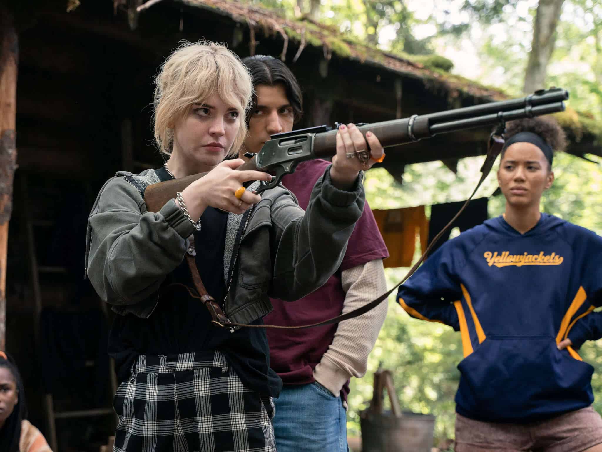 Natalie aiming a shotgun in the woods while Travis and Taissa look on