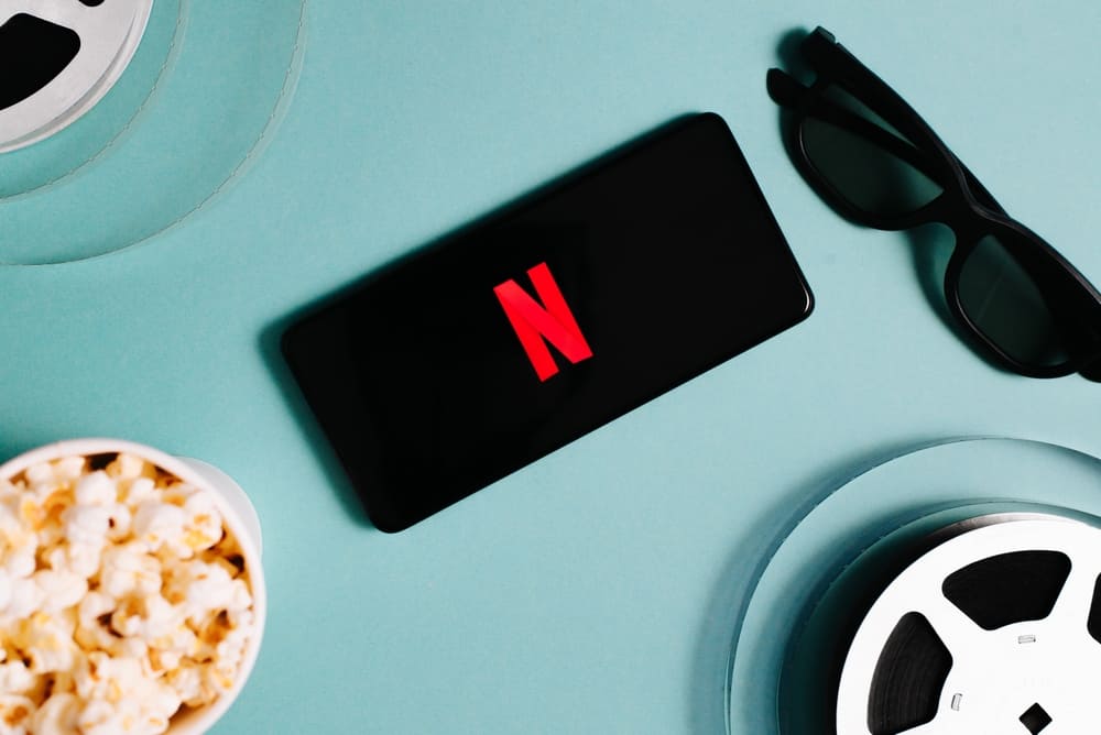 The Netflix logo appears on a phone screen against a light-blue backdrop. Two film reels, a cup of popcorn, and 3D glasses surround the phone.