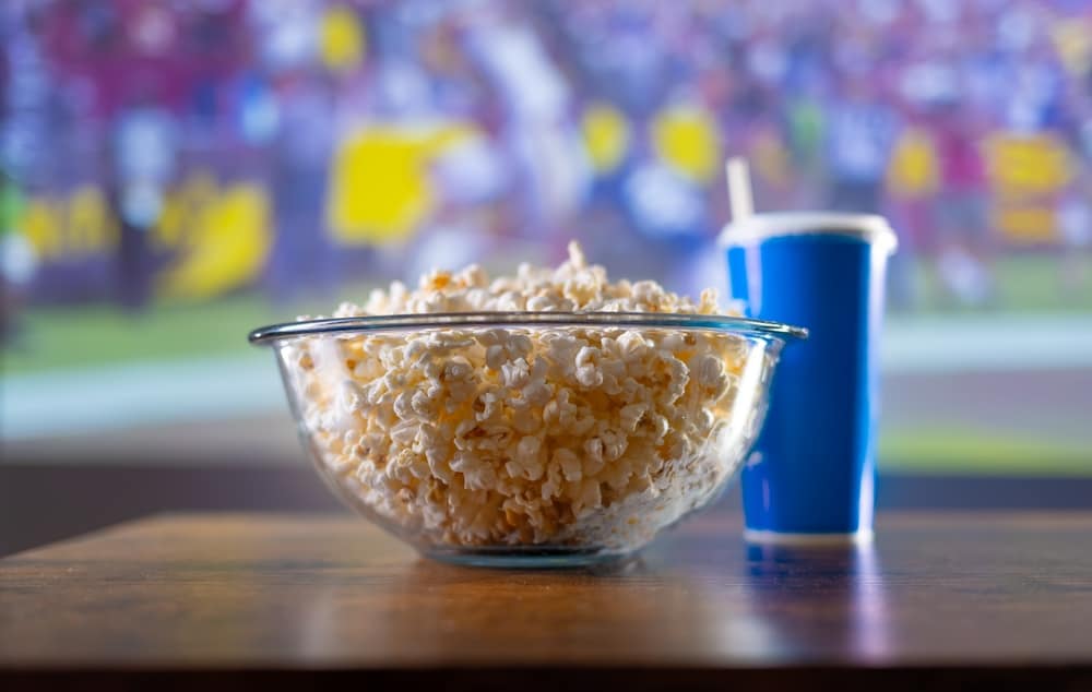 A glass bowl of popcorn and a plastic soda cup with a blurred TV in the background in this image from Shutterstock