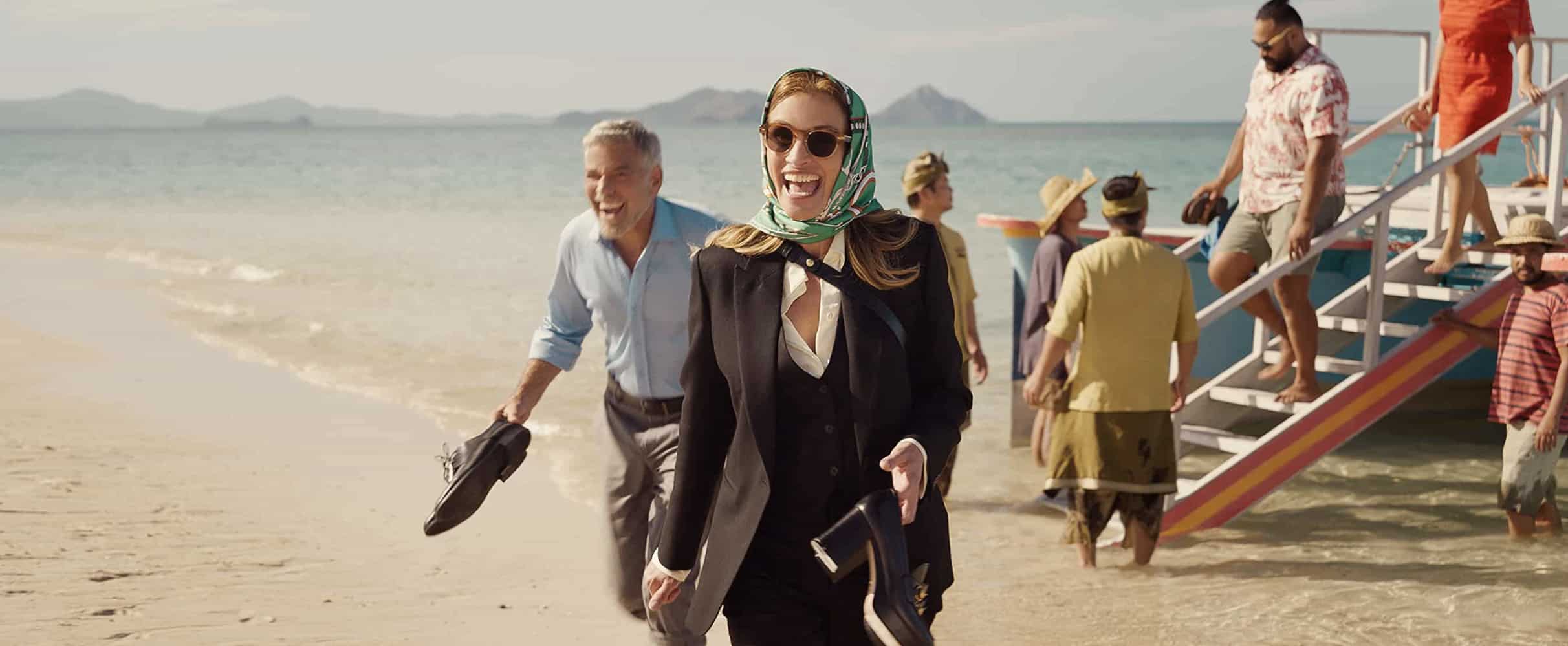  A woman with a big smile, a teal headwrap, and a black suit is in the forefront, and behind is a man in a blue button-down shirt in this image from Peacock.