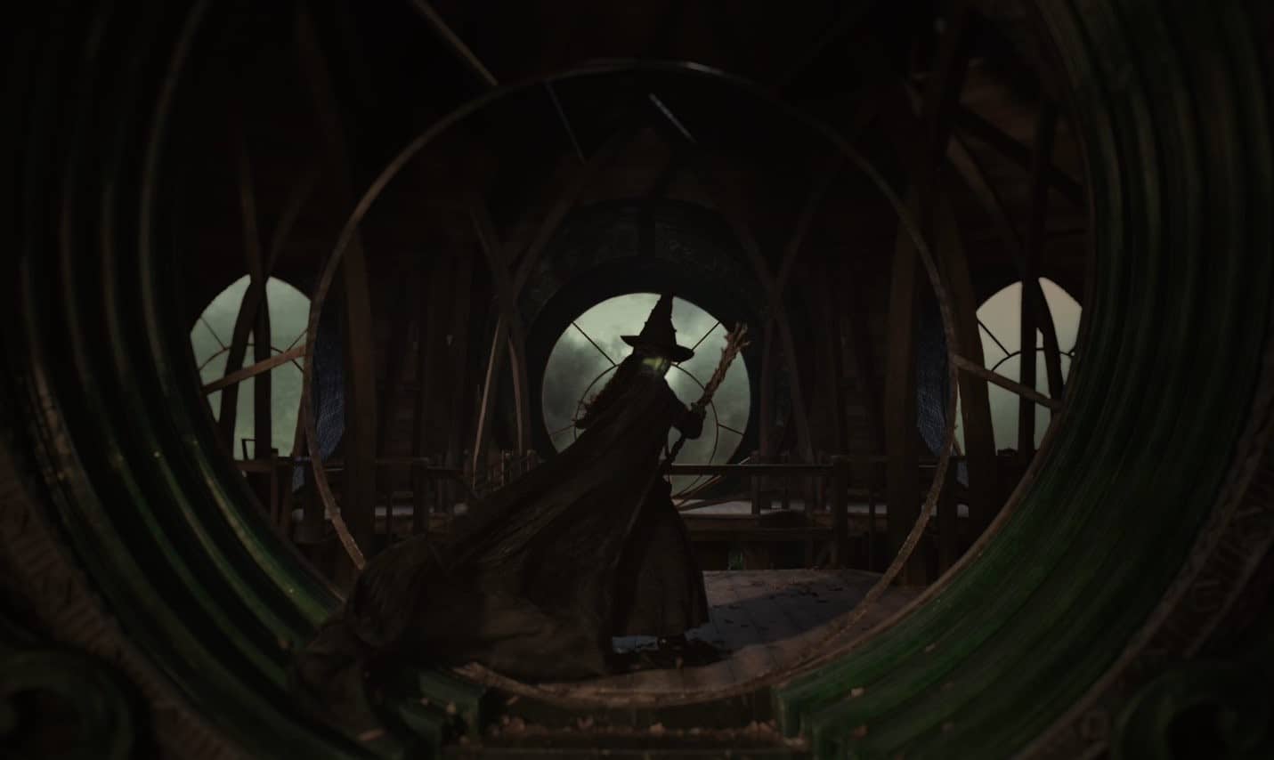 A woman in a witch’s cloak and hat is seen from behind carrying a broomstick in a clock tower in this image from Universal Pictures.