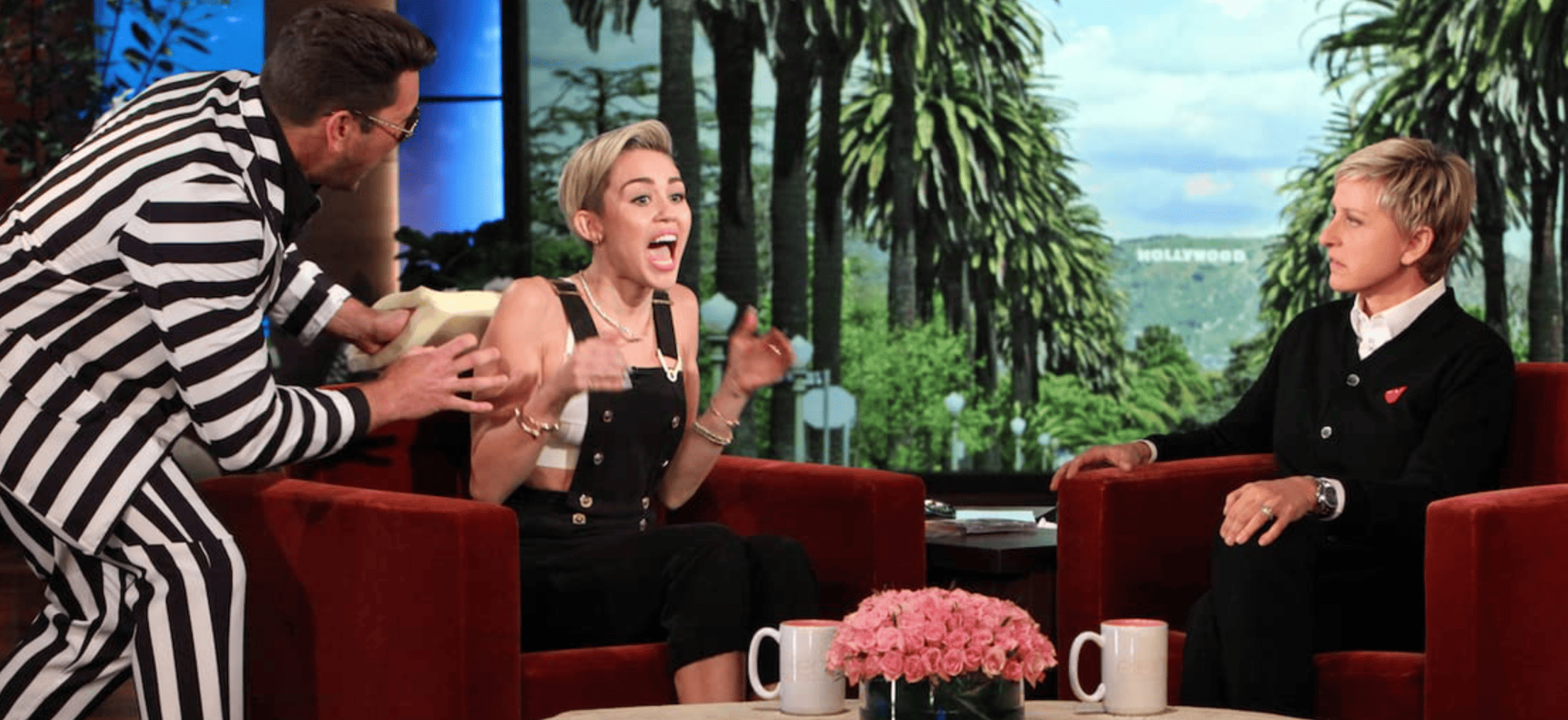 Ellen DeGeneres sitting on her signature couch next to a startled Miley Cyrus in this image from A Very Good Production