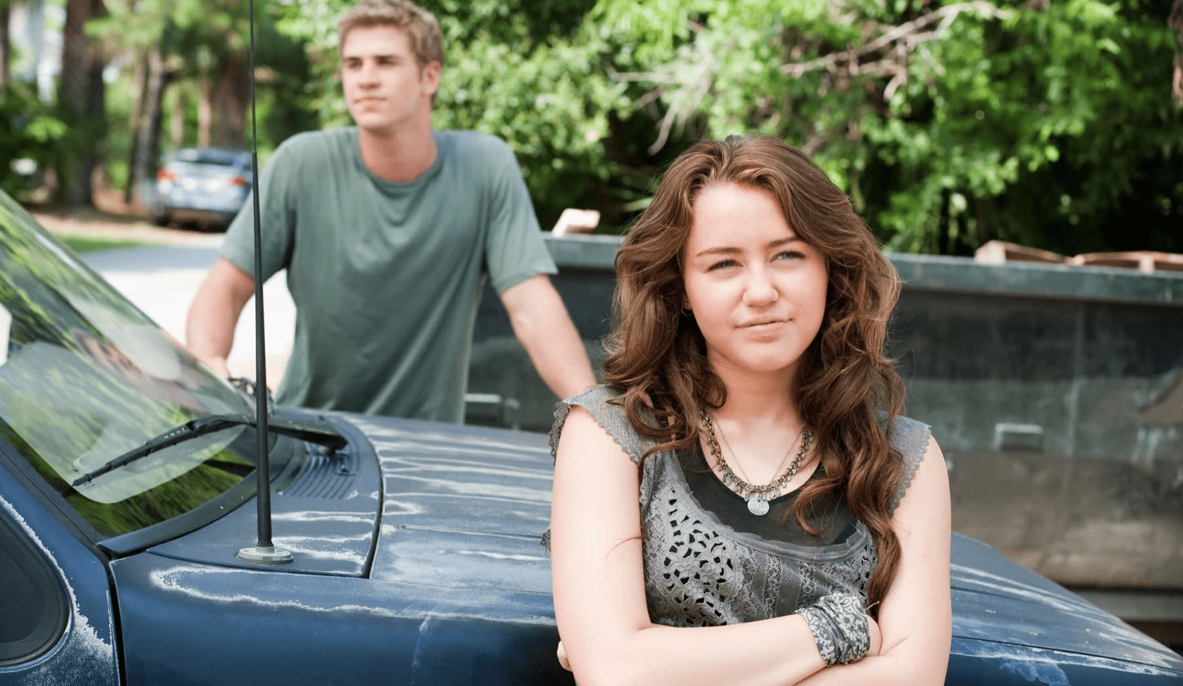 A young Miley Cyrus poses in front of a car while Liam Hemsworth hangs out in the background in this image from Walt Disney Studios.