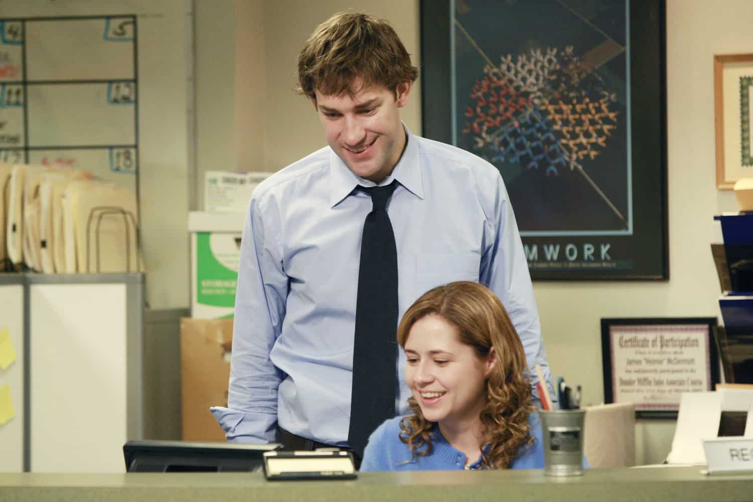 A businessman and a receptionist laugh at a computer together in this image from Peacock.