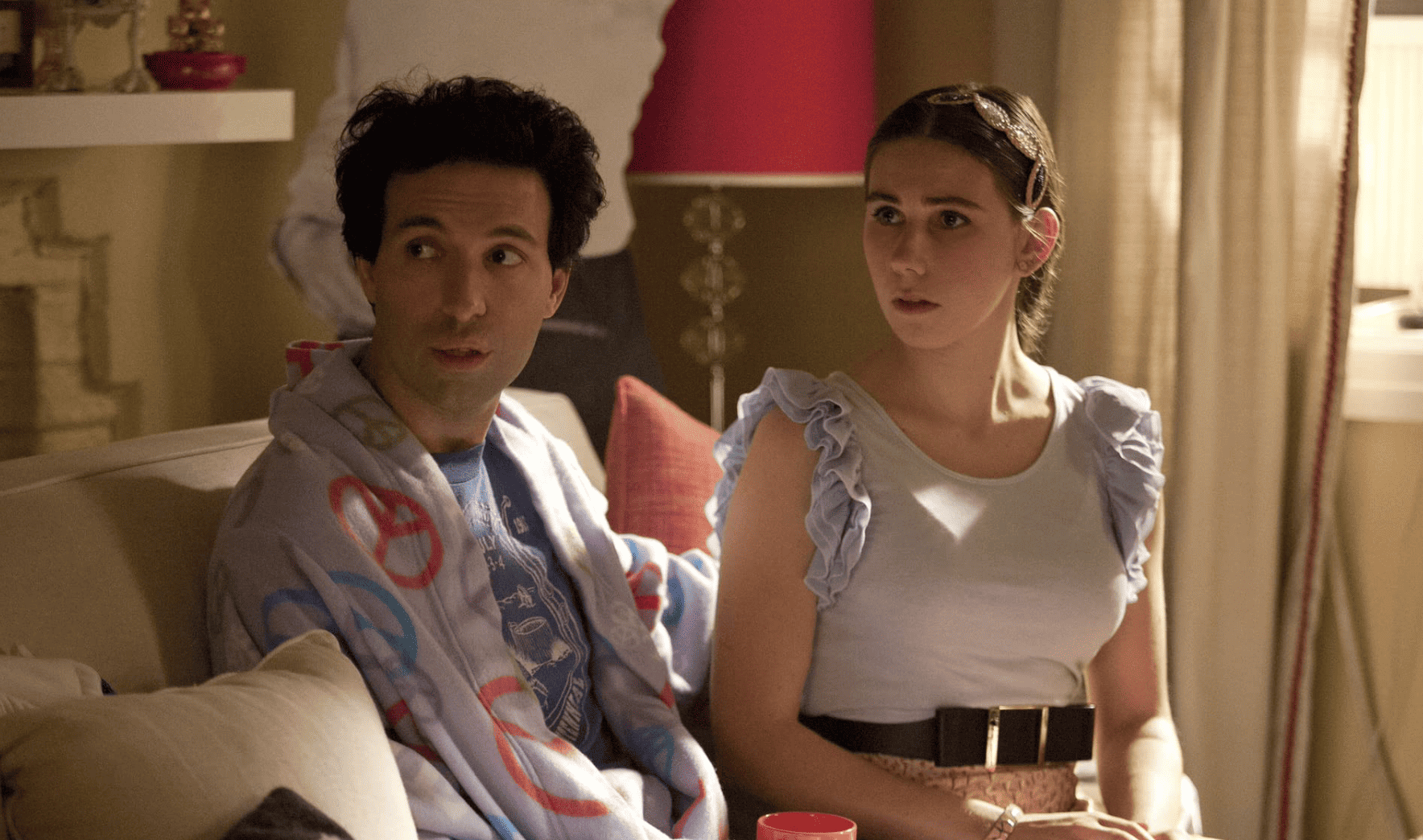 Shosh in her living room wearing a ruffled shirt and leopard-print skirt while her boyfriend Ray wears her peace-sign robe in this image from Apatow Productions