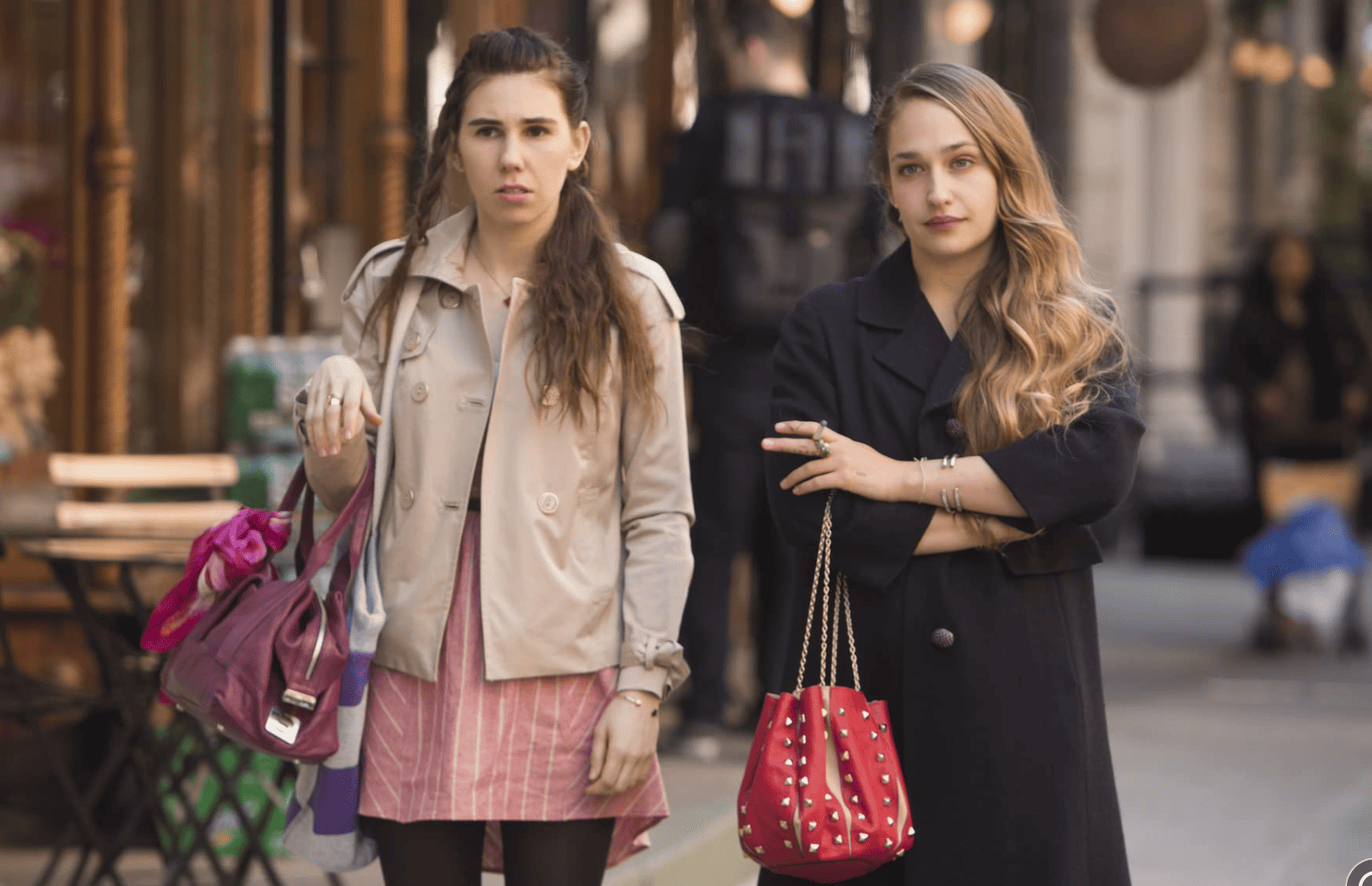 Shosh, with half of her hair in a braid, is standing in the middle of the street, holding a purse with a ribbon and standing next to Jessa in this image from Apatow Productions