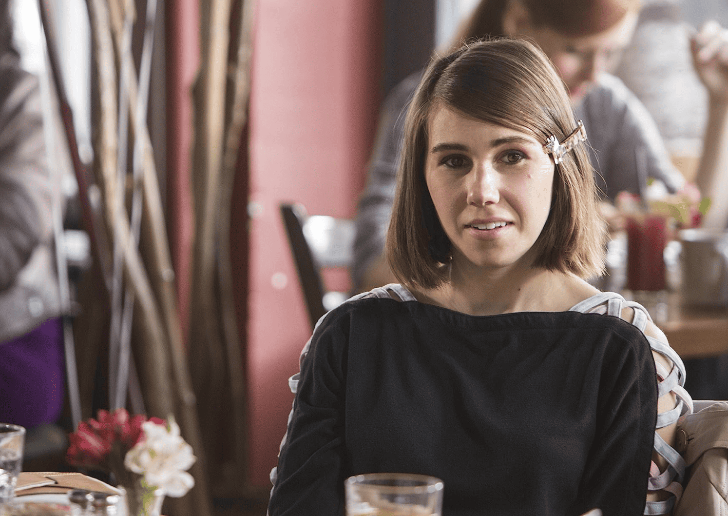 Shosh is sitting at a restaurant with a shoulder-length bob partially held by a barrette in this image from Apatow Productions.