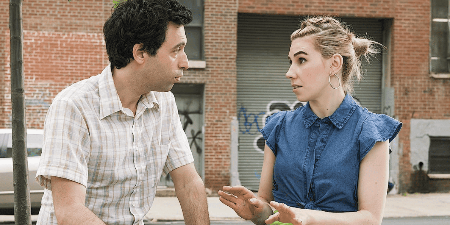 Shoshana is sitting on a bench wearing a denim sleeveless shirt and a bright green skirt next to her boyfriend Ray in this image from Apatow Productions