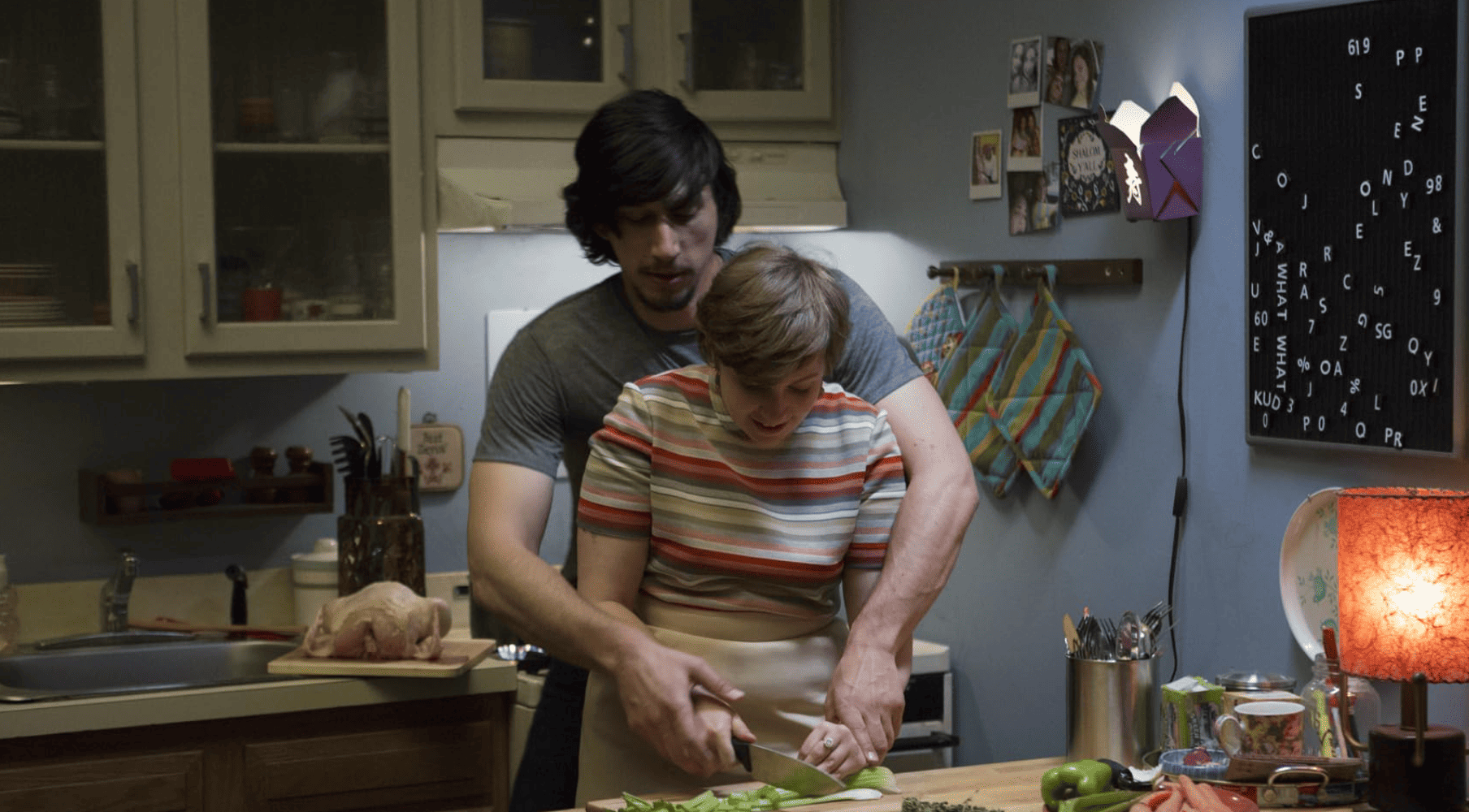 Adam Driver wrapping his arms around Lena Dunham from behind in a kitchen in this image from Apatow Productions