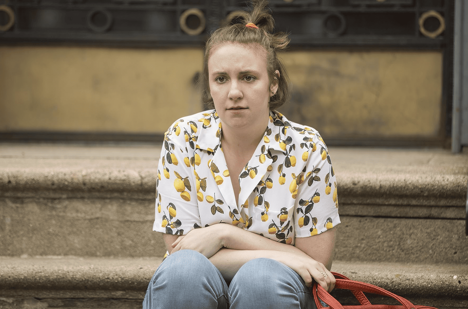 A visibly worried Lena Dunham sitting on some front steps with her legs crossed over her knees in this image from Apatow Productions