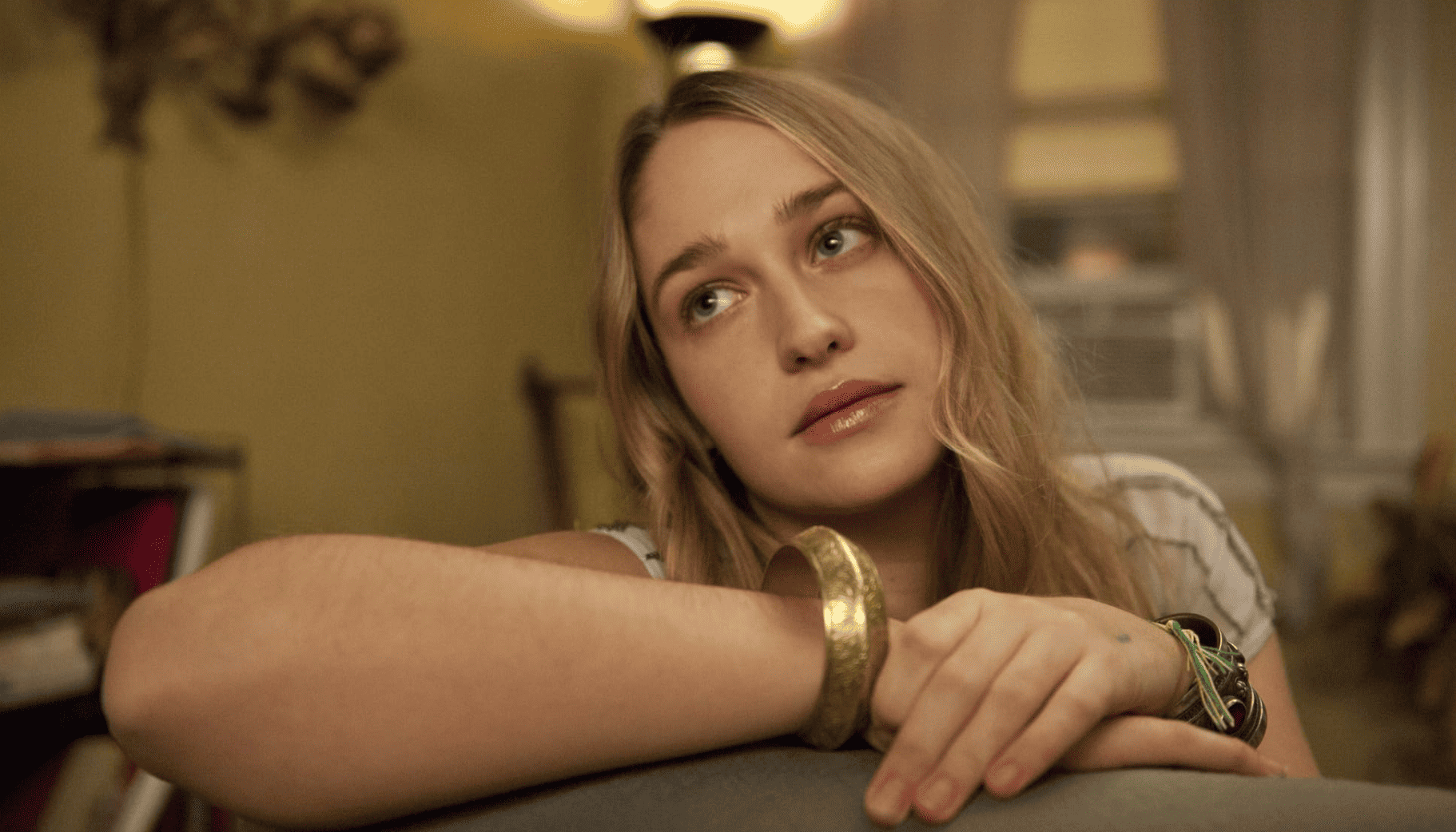 A wide-eyed Jemima Kirke resting her arms on the back of the couch while staring off-camera at the ceiling in this image from Apatow Productions