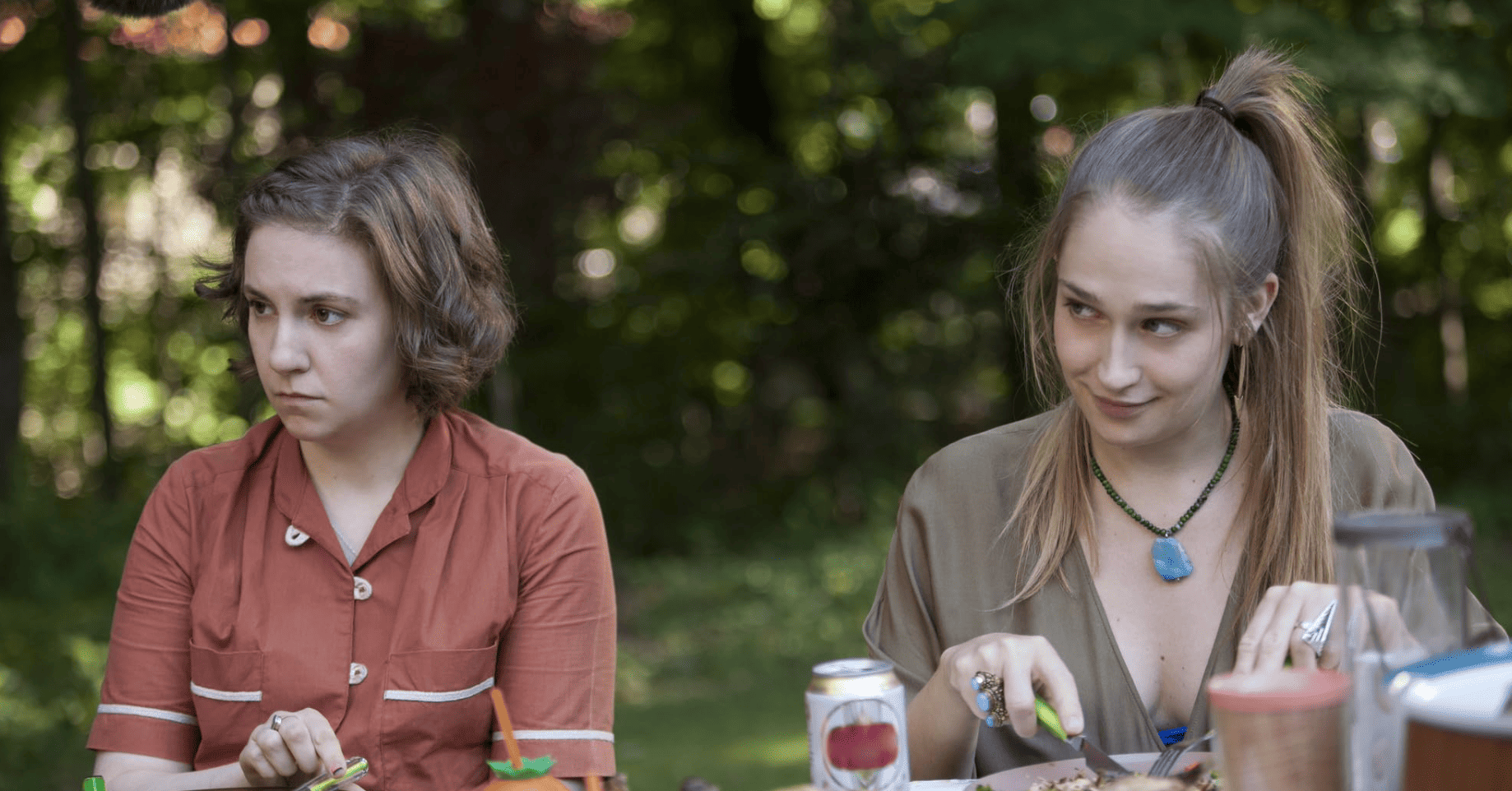 Jessa coyly looks off-camera while sitting at an outdoor dinner table next to Hannah eating rabbit in this image from Apatow Productions.