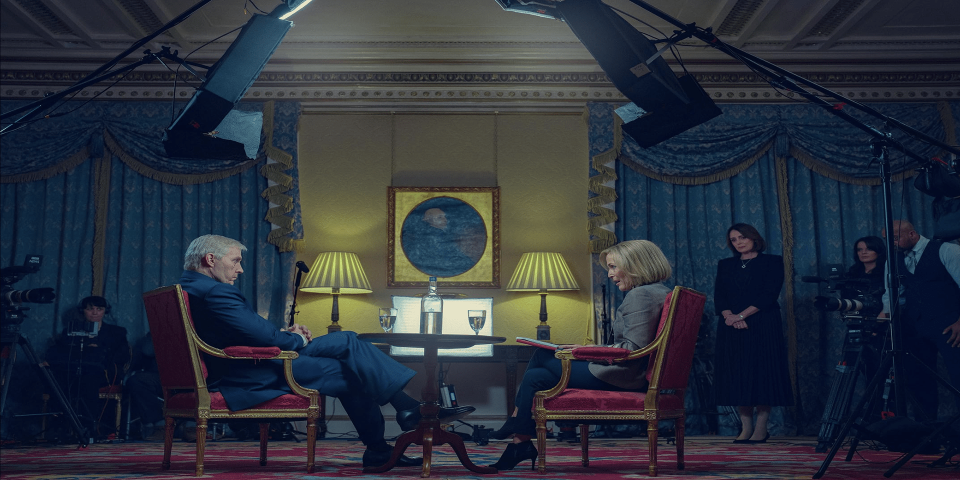 A man in a suit and a female reporter sit in a staged room to film an interview in this image from The Lighthouse.