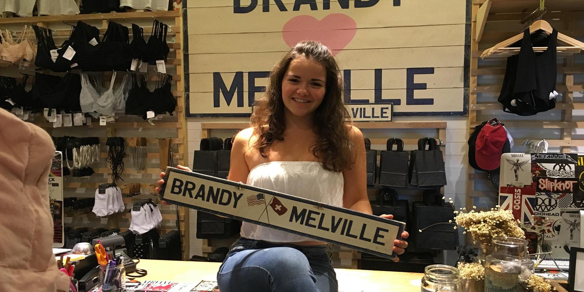 A young girl sits on a cashier table holding a Brandy Melville sign in this image from HBO Documentary Films.