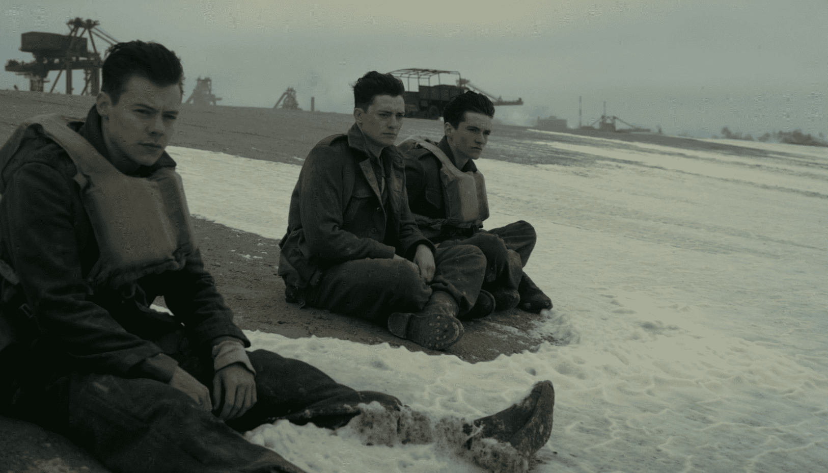 Harry Styles sitting in the foreground of a beach dressed in military uniform while two other soldiers sit farther back in this image from Warner Bros. Pictures