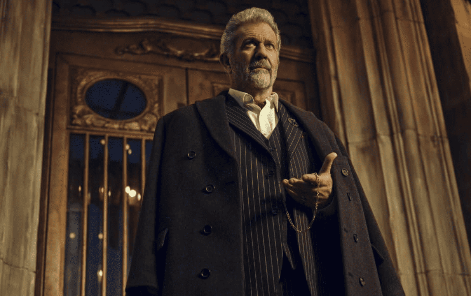 A man standing against the gilded doors of the hotel, sporting a full white beard and a pinstripe suit while holding a pocket watch in this image from Thunder Road Pictures.