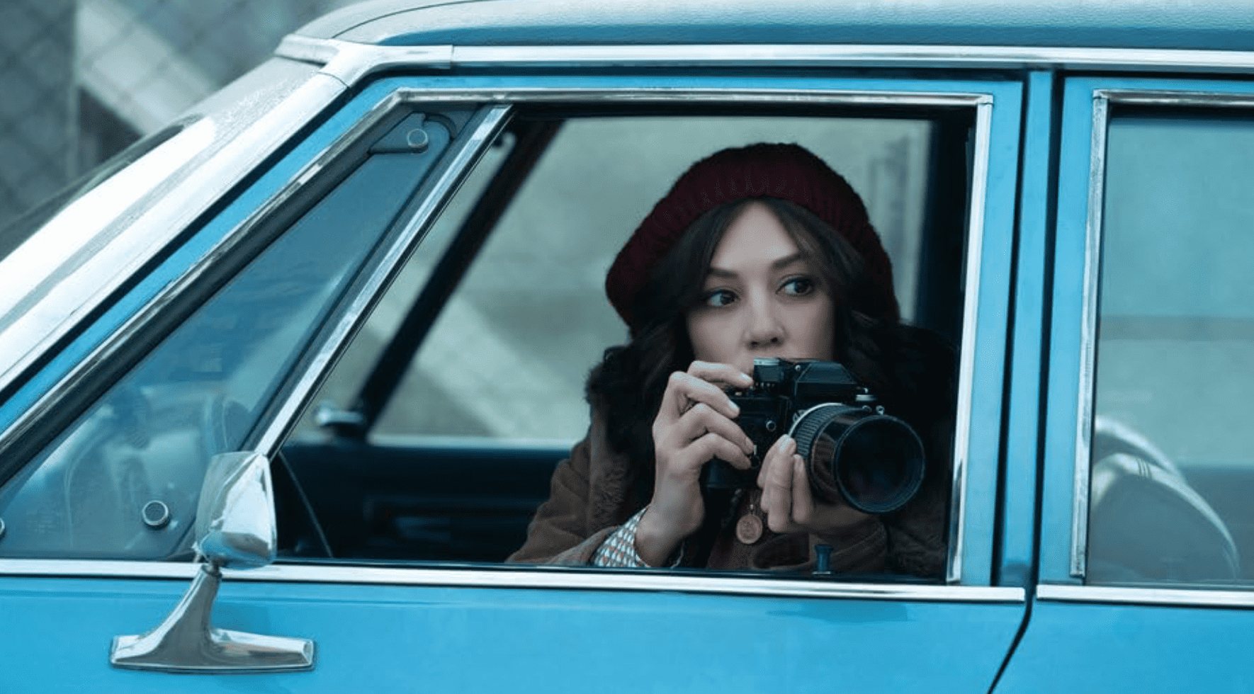 A woman leans out of a parked car holding a camera, presumably surveilling someone in this image from Thunder Road Pictures.