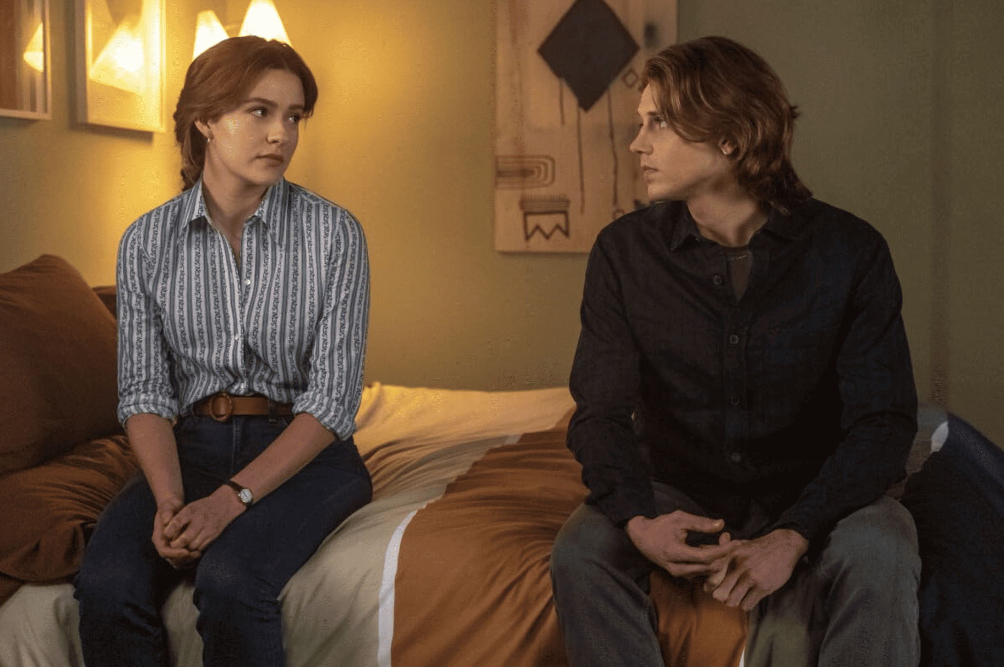 A young woman and man sit a few feet apart on a bed in this image from CBS Television Studios. 