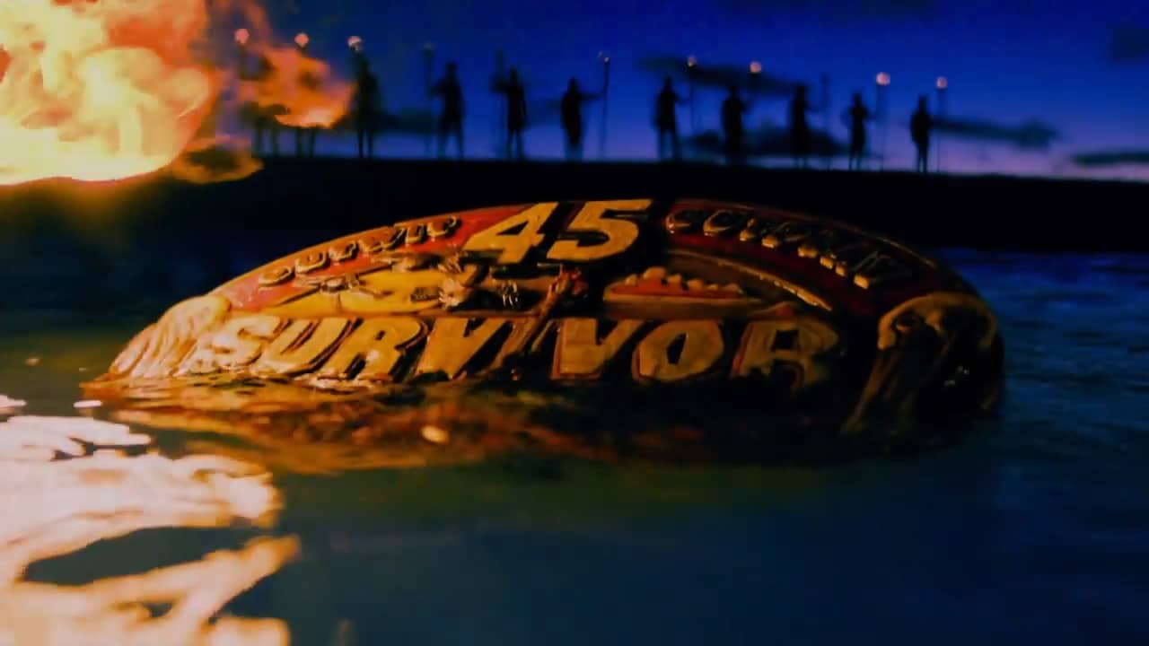 A piece of wood floats in the water with flames nearby, while a group of people in the background hold torches in this image from MGM Television.