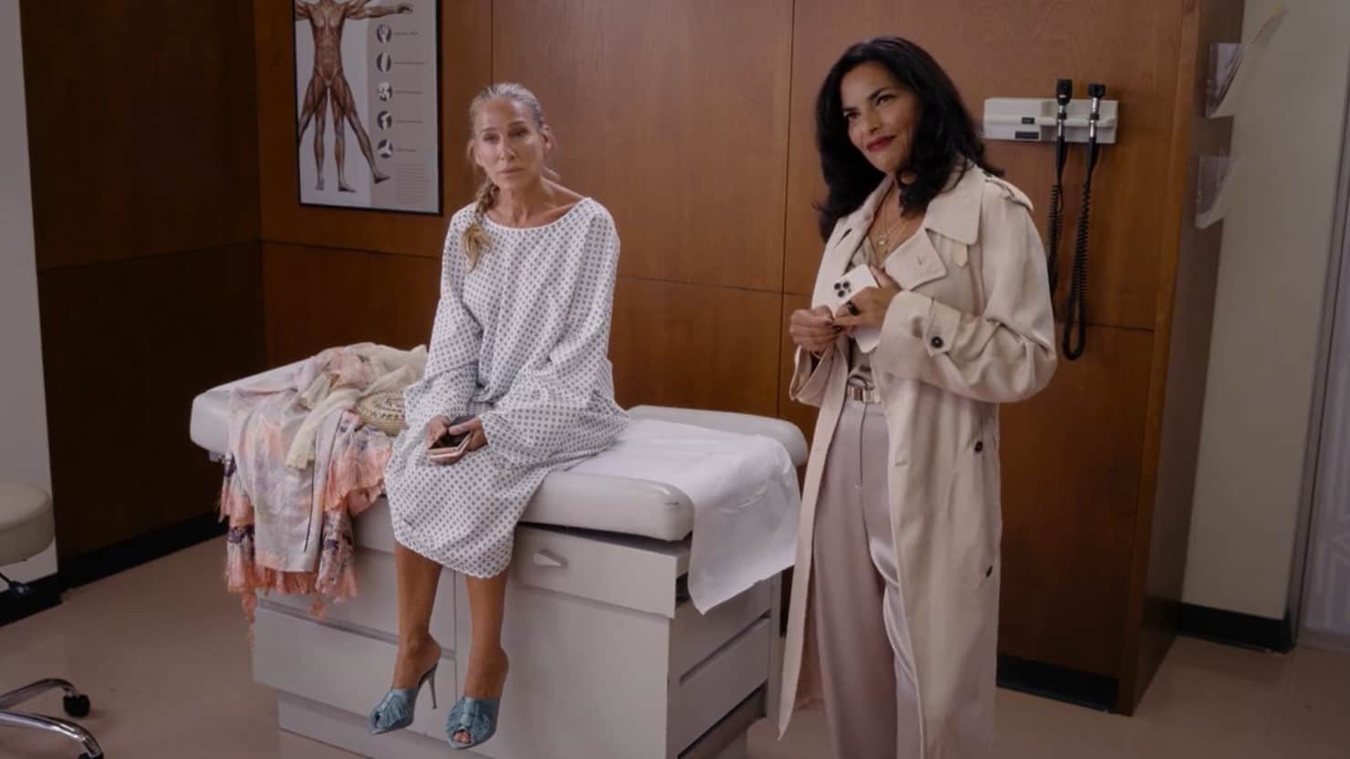 A woman wearing a hospital gown sits in a doctor’s office with a friend standing beside her in this image from HBO Entertainment.